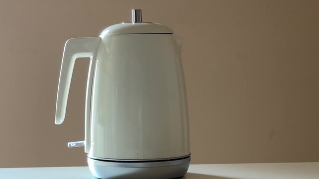 A Hand Turns On A White Modern Electric Kettle Standing On A White Table Against A Beige Wall In Soft Daylight 