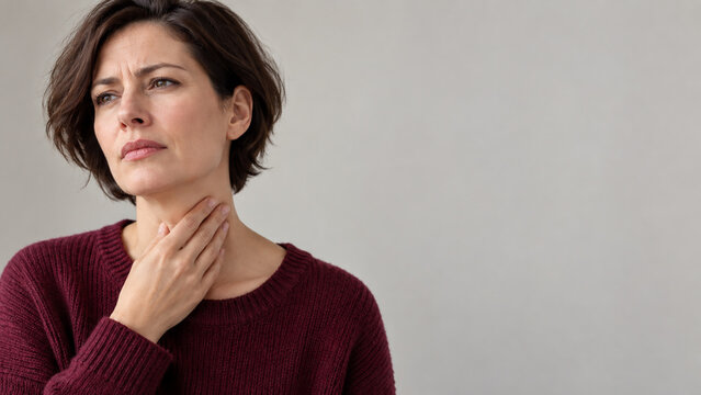 Middle-aged woman touching her painful throat with a concerned expression against a neutral background, illustrating discomfort associated with colds and upper respiratory infections.