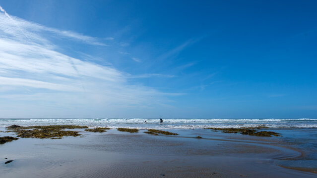 Vast beach in Cornwall with two lone surfers walking towards the sea, featuring large copy space in the sky