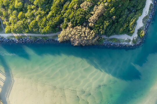 Aerial downward view of rippled sand beneath clear turquoise water in Back Creek. A deeper channel follows the rocky bank beside dense coastal vegetation and a walking path
