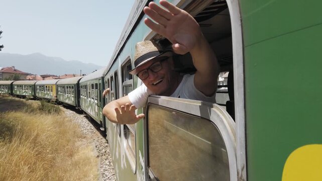 A man is waving from the window of a train open window traveling on track. The train is green and is traveling down the tracks