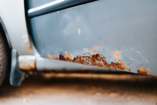 The lower side of a vehicle shows significant rust damage with visible corrosion and peeling paint. The background is blurred, emphasizing the wear on the metal surface