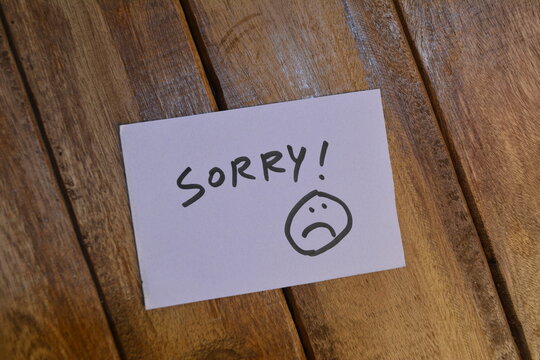 a handwritten note saying sorry with a sad face emoticon rests on a wooden surface, conveying a sentiment of apology