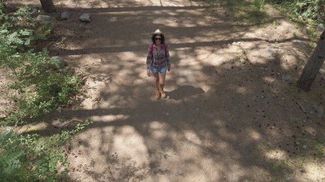 A young hiker girl is walking down a path in the woods. She is wearing a red backpack and a hat. The path is surrounded by trees and bushes, and the sunlight is shining through the leaves, aerial shot