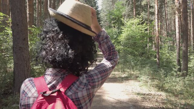 A woman with curly hair puting hat on head and walks down a path in the woods. She is wearing a red backpack and a plaid shirt
