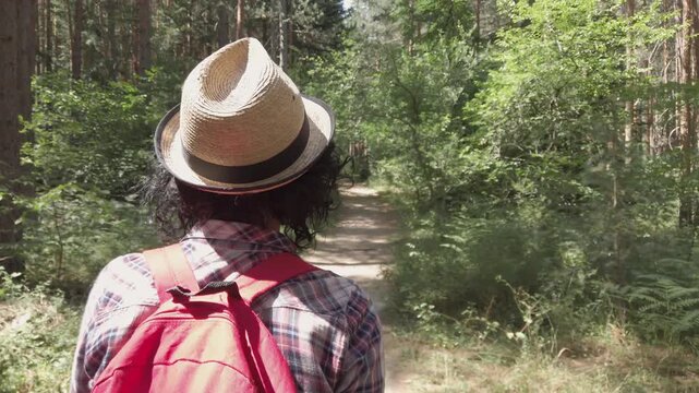 A woman with curly hair puting hat on head and walks down a path in the woods. She is wearing a red backpack and a plaid shirt