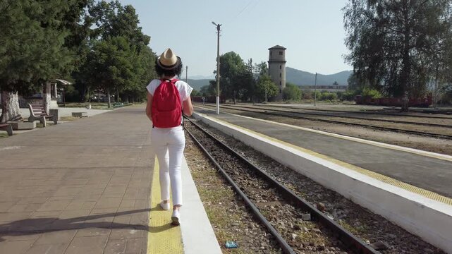 A woman wearing a straw hat and a red backpack is standing on a train platform. She is smiling and looking at the camera