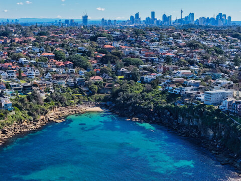 Aerial view of Gordons Bay with turquoise waters, rocky cliffs, and surrounding residential houses, with the Sydney skyline in the background, Coogee, New South Wales, Australia.