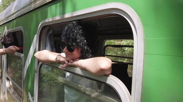 Traveler woman is leaning on a train window with her head resting on her hand. She is wearing sunglasses and a green shirt