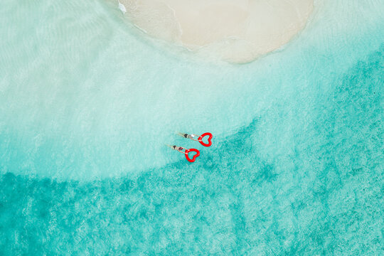 Aerial view of two people swimming in crystal clear turquoise water with red heart-shaped floats near a white sand beach in Vashafaru, Haa Alif Atoll, Maldives.
