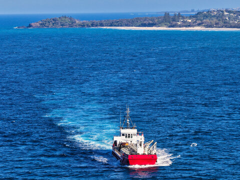 Aerial view of a red work boat navigating the deep blue waters near Cook Island, leaving a white wake behind, with the coastline of New South Wales, Australia.