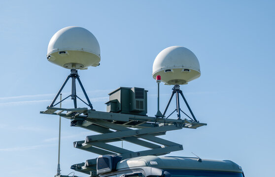 UAV ground control station part: two white radome satellite antennas on tripod mounts atop a raised scissor lift mast, green electronics cabinet and red warning light, clear blue sky