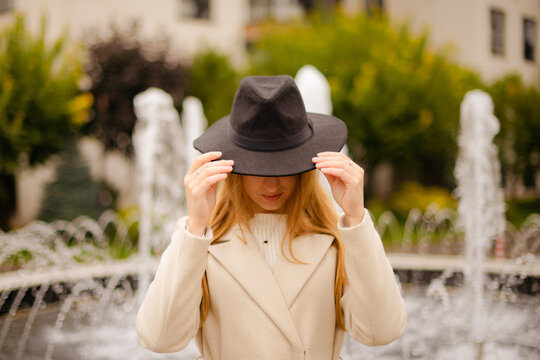 woman in beige coat lowering black hat brim over face in fall park near fountain