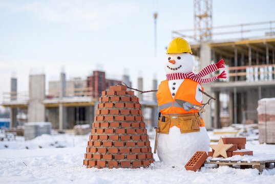 Snowman bricklayer in a hard hat and safety vest building a small conical brick tower with a trowel, festive winter construction site with snow, pallets and a wooden star ornament