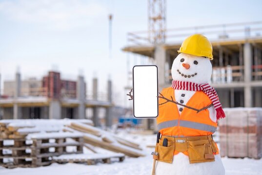 Snowman builder in a yellow hard hat, orange safety vest and tool belt holding a blank white smartphone screen for mockup, winter construction site with crane and unfinished building in the background