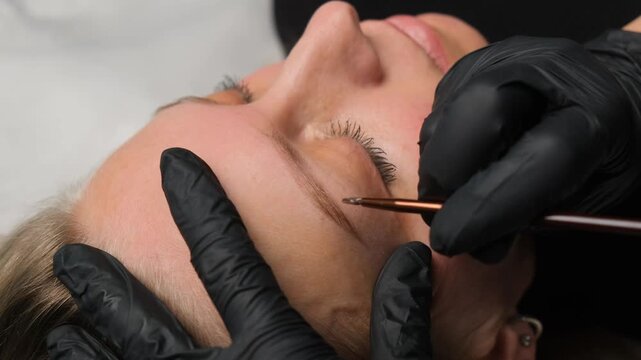 Professional cosmetologist performing permanent makeup procedure on a woman's eyebrows in a salon