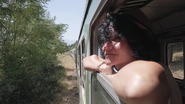 Female wearing a hat is sitting on traveling train window and looking at landscape on sunny summer day