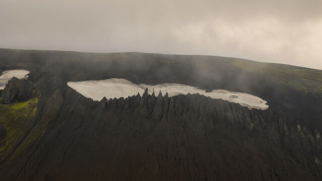 Aerial view of Landmannalaugar volcanic landscape featuring dark rock formations, patches of snow, and misty clouds in Hella, Rangarthing ytra, Iceland.