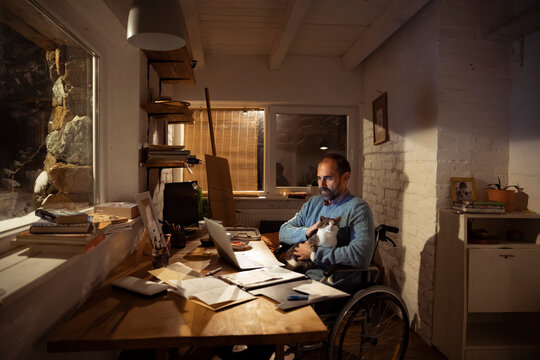 Man in wheelchair using laptop with cat in home office