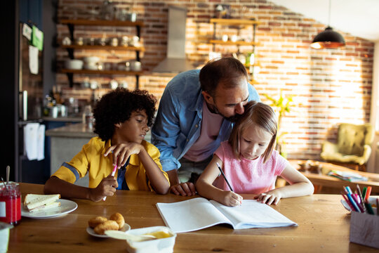 Man helping girl write at kitchen table while boy watches