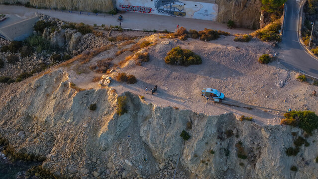 Aerial view of a rocky coastal cliff with a parked blue van and people standing nearby, overlooking the Mediterranean sea in Alicante, Valencian Community, Spain.