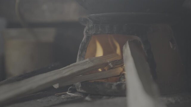 Log Profile Handheld close up of firewood burning inside a traditional clay stove in a rustic village kitchen, with warm orange flames glowing through the opening. 
