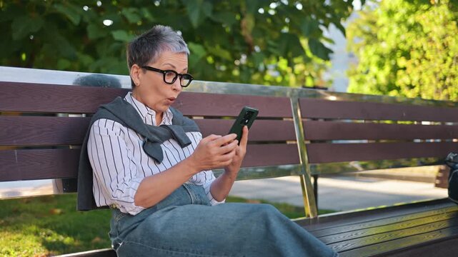 Mature woman with gray hair and glasses sits on a bench, focused on her smartphone, with parked cars and plants visible in the outdoor setting