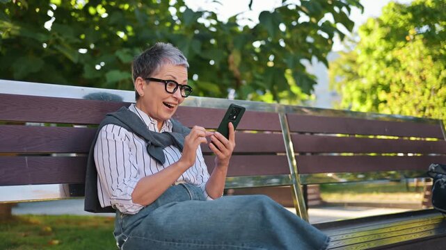 Mature woman with gray hair and glasses checks her smartphone while seated on a bench in an urban area, surrounded by parked cars and buildings