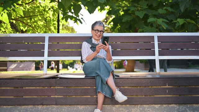 Mature woman with gray hair and glasses sits on a bench, focused on her smartphone, with parked cars and plants visible in the outdoor setting