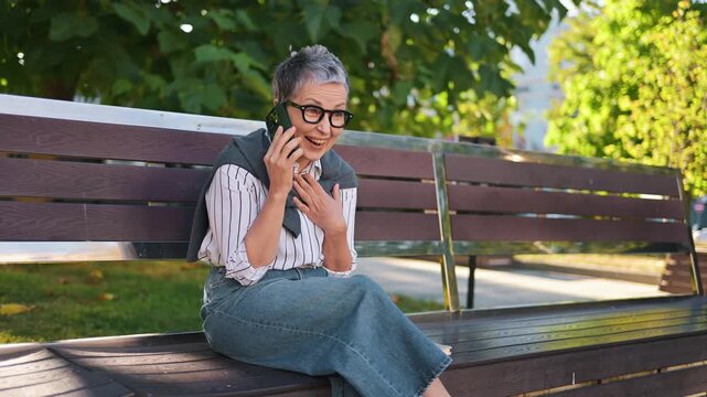 Senior woman with gray hair and glasses engages in a phone conversation while seated on a bench in a sunny urban park with cars visible in the background