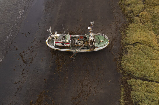 Aerial view of an old abandoned fishing boat stranded on the dark sand of Rif Beach, surrounded by patches of green grass and the calm sea in Rif, Iceland.