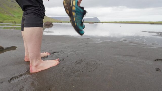 A barefoot hiker carries their shoes while crossing a muddy tidal flat in the remote Hornstrandir Nature Reserve on Iceland. Lush green hills and cliffs frame the serene Westfjords landscape.