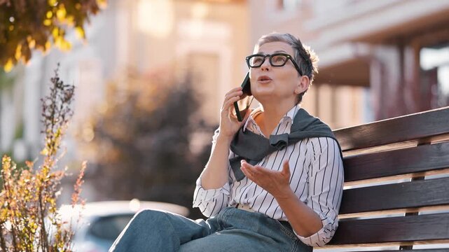 Senior woman with gray hair and glasses engages in a phone conversation while seated on a wooden bench in an urban environment with greenery
