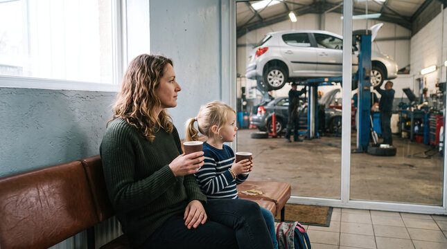 Mother and daughter sitting in a garage waiting area while mechanics repair their car.
