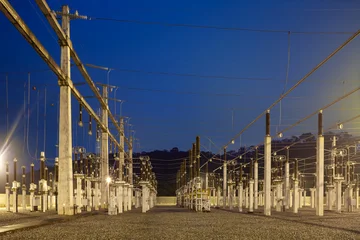 An electrical substation in Brazil is illuminated at dusk, with rows of insulators, overhead conductors and concrete supports forming a dense network above a gravel yard. © Jose Luis Stephens