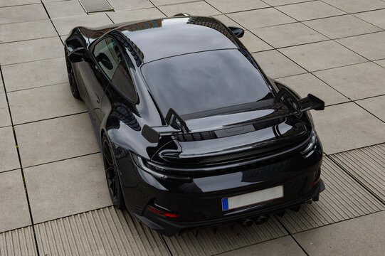 A high-angle rear view of a black high-performance sports car parked on a modern paved plaza. The photograph highlights the vehicle&rsquo;s wide body, large aerodynamic rear wing, sculpted rear bodywork.