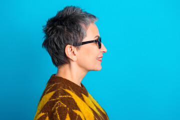 Smiling mature woman in profile with short gray hair and glasses against bright blue background © deagreez