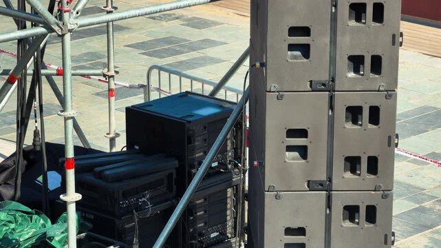 A line array speaker system stacked beside metal scaffolding and audio equipment cases on an outdoor stage, concert setup on a city square before an open-air event. High quality 4k footage