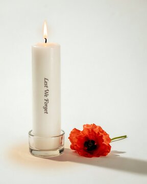 Lit white memorial candle with lest we forget text and red poppy flower on white surface.