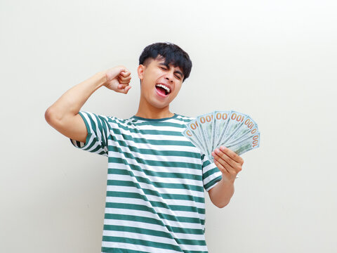 An excited young Asian man holding money fan and celebrating success against studio background for financial loan and online scholarship reward campaign