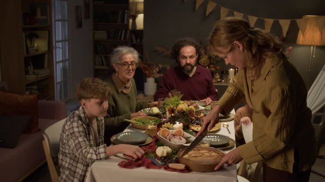 Middle aged Caucasian woman slicing pumpkin pie and serving slice to teenager during festive family holiday dinner. Perfect for Thanksgiving campaigns and autumn food marketing