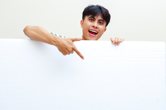 Young Southeast Asian man pointing down at blank white board happy excited face against plain background ideal for commercial promotion advertising copy space