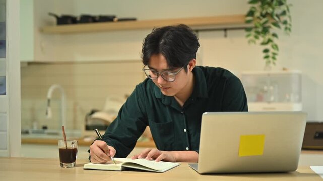 Asian Man Writing Notes in Notebook While Working from Home