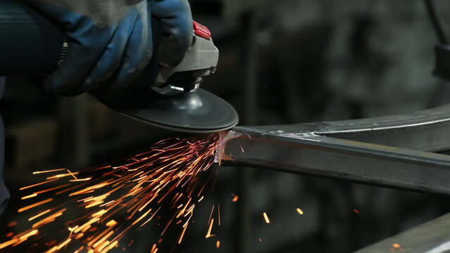 Worker uses angle grinder to shape steel component in plant workshop closeup. Hot sparks scatter across frame. Metalworking industry concept