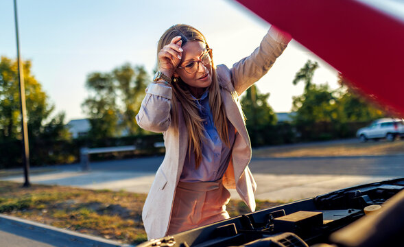 Worried business woman looking under open hood of broken car.
