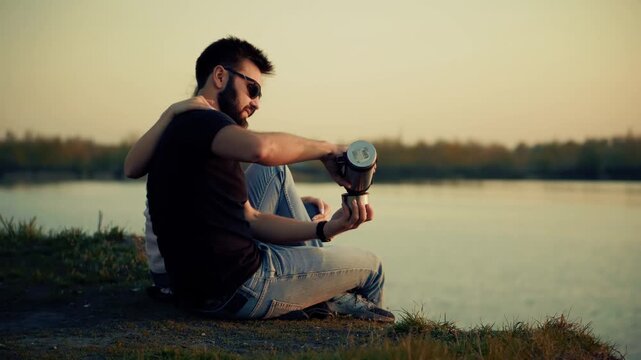Couple Sitting On Lake Shore At Sunset And  Pouring Warm Drink From Thermos Mug During Outdoor 
