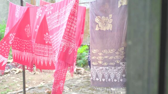 Pink and purple batik fabrics with fringed edges hanging on a clothesline and fluttering in strong wind outdoors, traditional Indonesian batik textile drying after dyeing