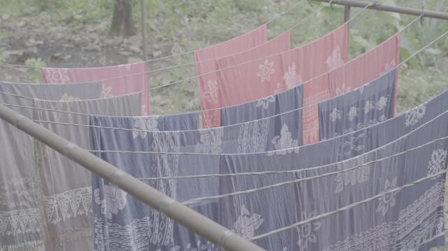 Ungraded log footage of colorful batik fabrics in pink and purple hanging to dry on wire lines and a metal frame outdoors with green foliage in the background, traditional Indonesian batik textile