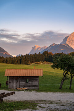 Traditional wooden alpine hut in a green mountain meadow with the rocky peaks of the Karwendel mountains illuminated by sunset light.
