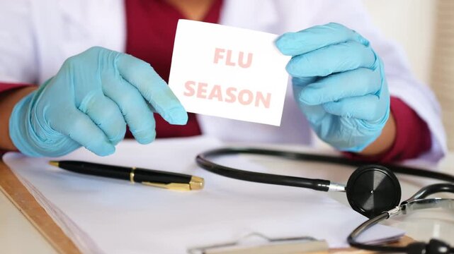 A doctor holds a sign reading "FLU SEASON." A transitional weather period. Pandemic, crisis, emergency situation. A warning about the season when the flu virus is prone to striking.
